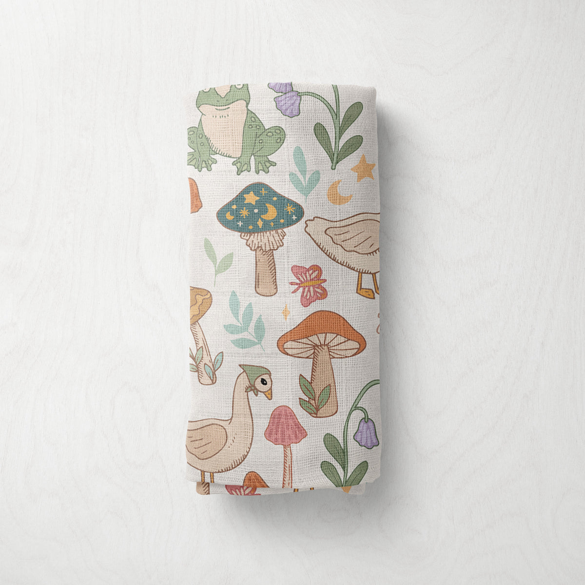 A rolled muslin swaddle, featuring a whimsical woodland pattern with mushrooms, birds, flowers, and leaves on a light cream background, displayed against a textured white wall.
