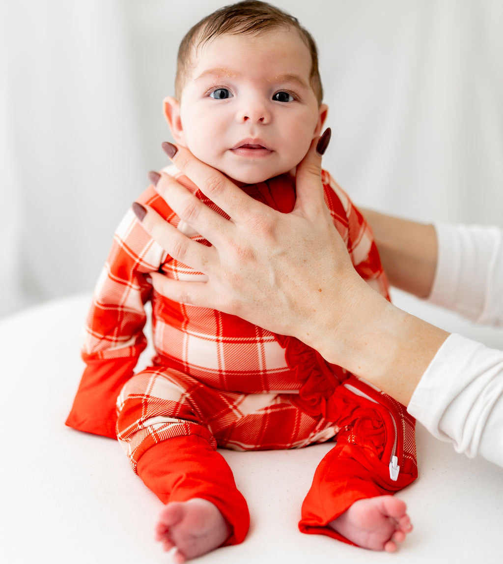 Baby in a red plaid outfit being held by an adult against a white background