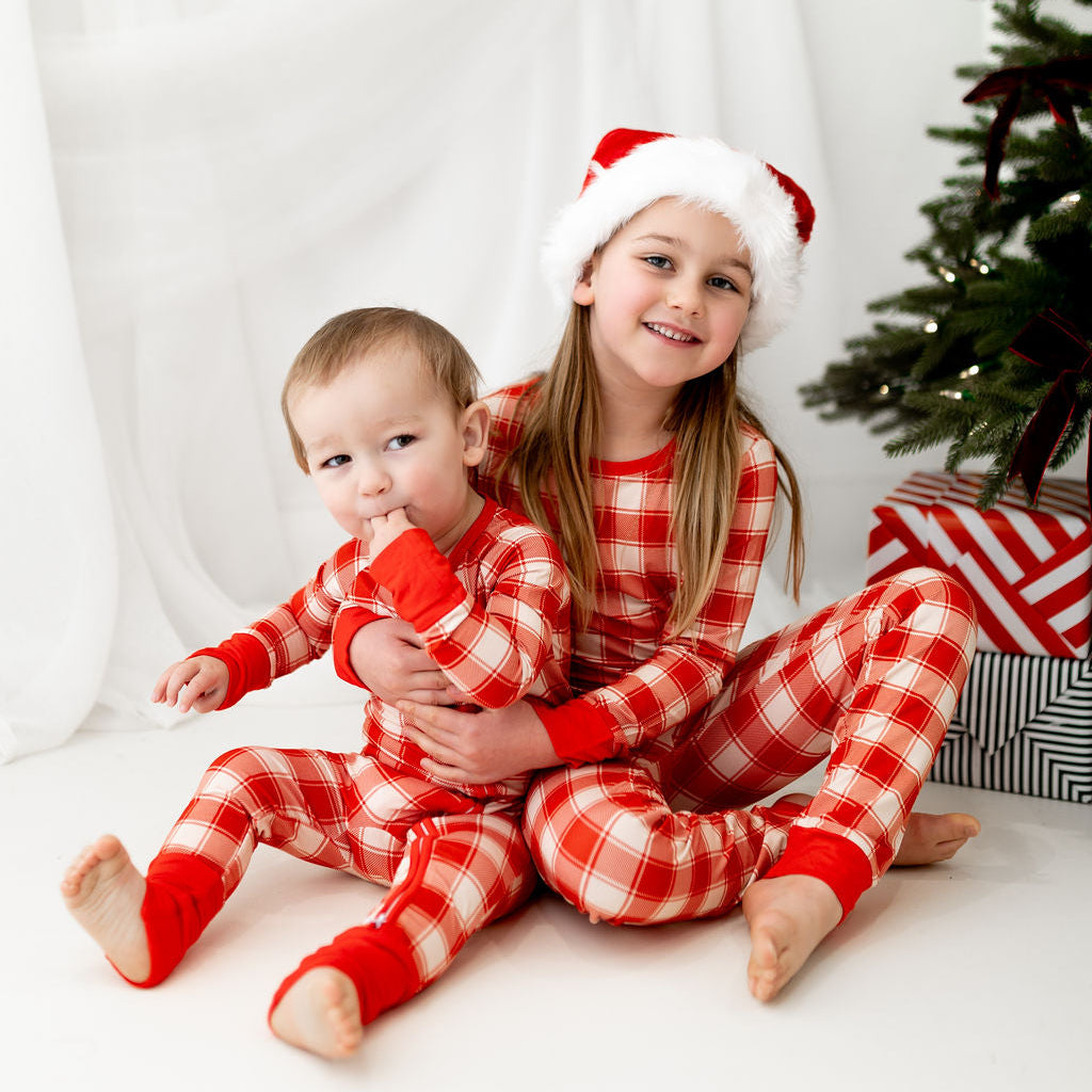 Two children in red and white checkered pajamas with Santa hats sitting in front of a Christmas tree.
