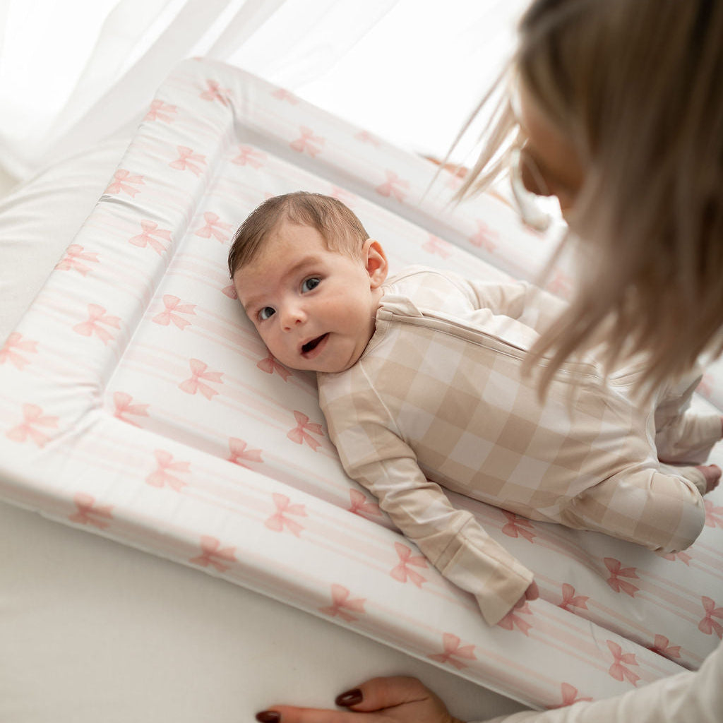 Baby lying on a white mat with pink patterns, being watched by a person.